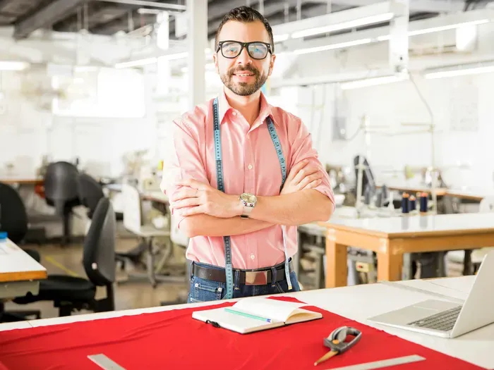 Confident tailor with glasses and measuring tape standing at a cutting table in a bright textile workshop